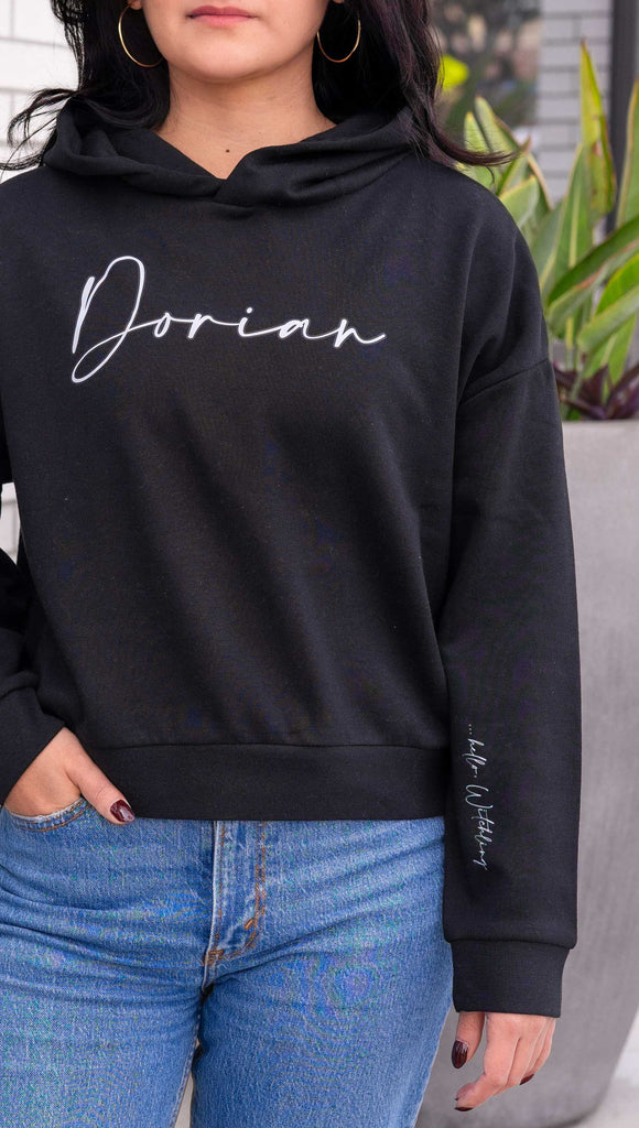 Model wearing WERKSHOP "Dorian" Hoodie. Officially licensed for Sarah J Maas. The hoodie says, "Dorian" on the chest and "... hello, witchling" on the wearer's left sleeve. Shown in Black with white writing.