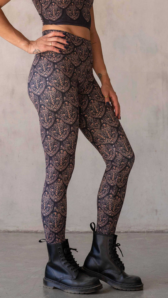 Side view of model wearing WERKSHOP Dragon Rider leggings in Gold. The artwork features an intricate battle shield designed to look like dragon scales. This color way is all shades of gold and bronze.