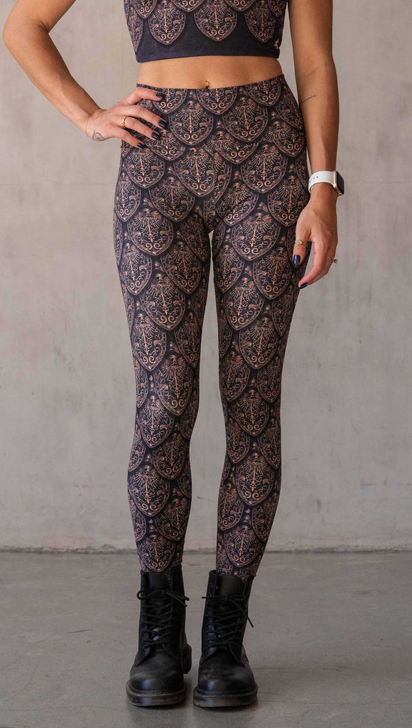 Front view of model wearing WERKSHOP Dragon Rider leggings in Gold. The artwork features an intricate battle shield designed to look like dragon scales. This color way is all shades of gold and bronze.