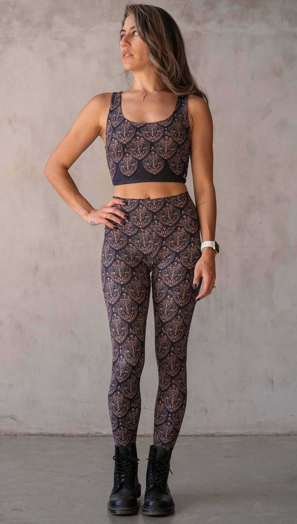 Front view of model wearing WERKSHOP Dragon Rider leggings in Gold. The artwork features an intricate battle shield designed to look like dragon scales. This color way is all shades of gold and bronze.