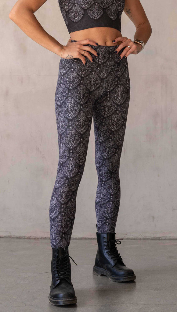 Front view of model wearing WERKSHOP Dragon Rider leggings in Silver. The artwork features an intricate battle shield designed to look like dragon scales. This color way is all shades of black and gray.