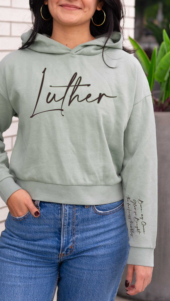 Model wearing WERKSHOP  "Luther" Boyfriend Hoodie. Officially licensed from Spark of the Everflame by Penn Cole. The hoodie has "Luther" on the front chest
... and "Burn, my Queen. Glow so Bright, the darkness trembles" on the sleeve. Shown here in Sage with dark taupe writing.