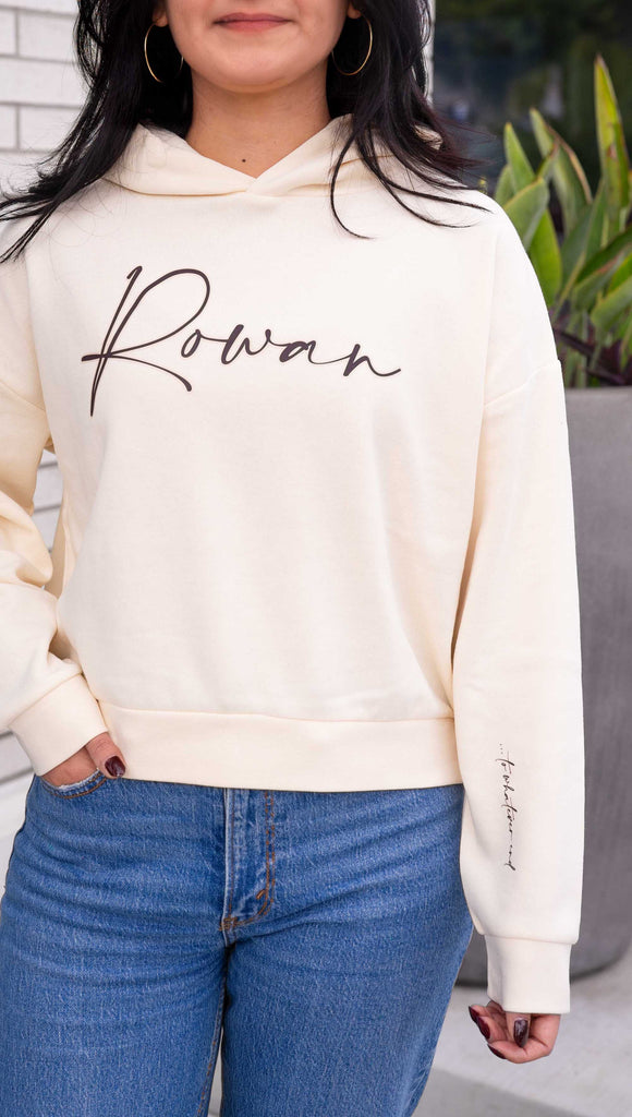 Model wearing WERKSHOP "Rowan" Hoodie. Officially licensed for Sarah J Maas. The hoodie says, "Rowan" on the chest and "... to whatever end" on the wearer's left sleeve. Shown in Bone with dark taupe writing.