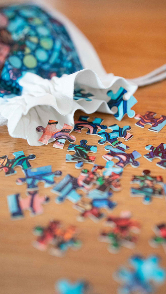 Jana Rose Puzzle puzzle pieces tumbling from the matching pouch. By Scott Christian Sava. The artwork on the puzzle features a woman with a wreath crown on her head and a bright red dress standing in front of a stained glass window.