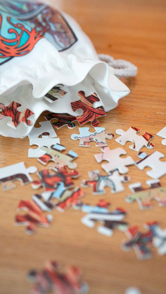 Art Nouveau puzzle pieces tumbling from a matching drawcord pouch. By Scott Christian Sava. The painting features a woman wearing a crown and pearls with long flowing red locks; standing in front of a round window.