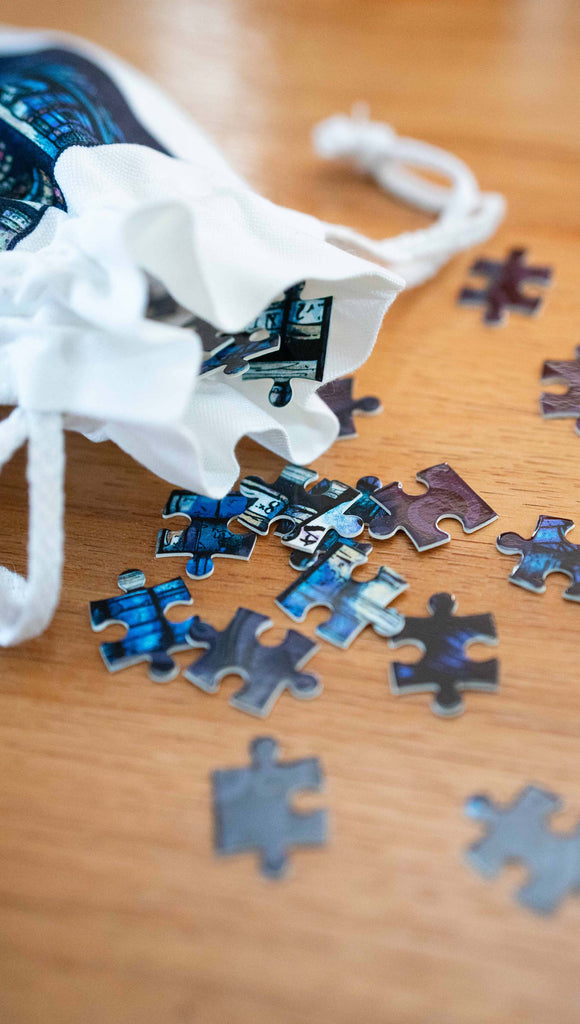 252pc jigsaw puzzle featuring a "Gothic Vampire" painting by Scott Christian Sava. The colors of the puzzle are very dark with pops of red. over grays and blues. This photo shows loose puzzle pieces tumbling from a matching drawcord pouch.