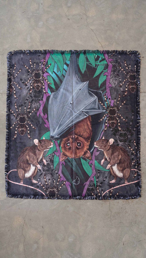 Decorative Chenille Tapestry printed with limited edition artwork by our Female Founder, Chriztina Marie. The artwork features an adorable fruit bat dangling upside down inside a tropical scene with a purple wreath of thorns. Under the bat, there are two rats. The background is a distressed dark gray brushstroke texture.