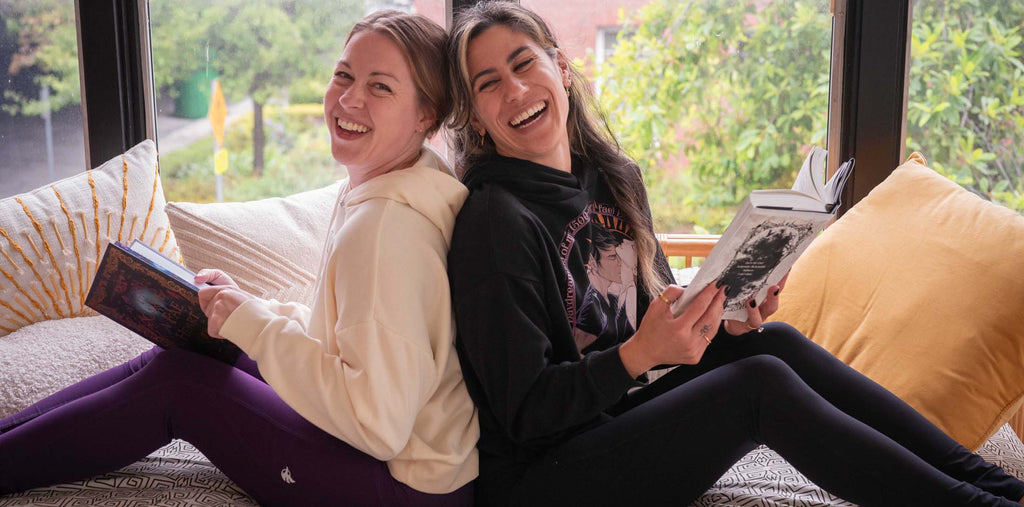 Two girls casually sitting back-to-back on a couch while reading and laughing. They're both wearing WERKSHOP Book Club hoodies and EnviSoft leggings