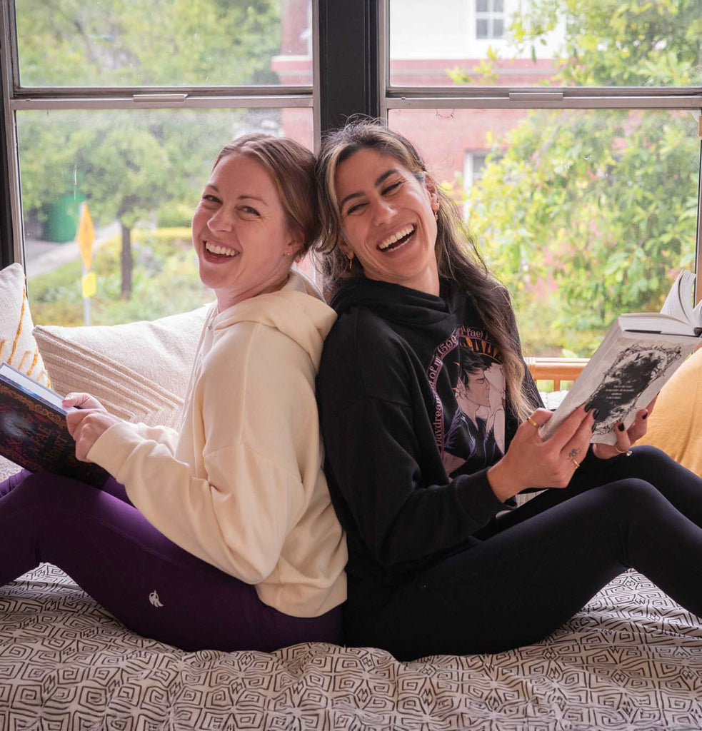 Two girls casually sitting back-to-back on a couch while reading and laughing. They're both wearing WERKSHOP Book Club hoodies and EnviSoft leggings