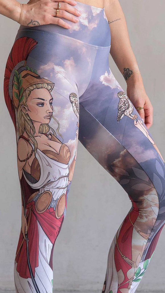 Zoomed in view of model wearing WERKSHOP Athena leggings. The leggings are printed with original artwork by Chriztina Marie and features Athena, the goddess of war standing on a cliff’s edge. She is holding a spear with one hand and her owl with the other. She is wearing a greek goddess dress/warrior hat with colors of cream, gold and red. Behind her is a beautiful cloudy sky with rays of light shining down onto her. The model is also wearing a matching top.