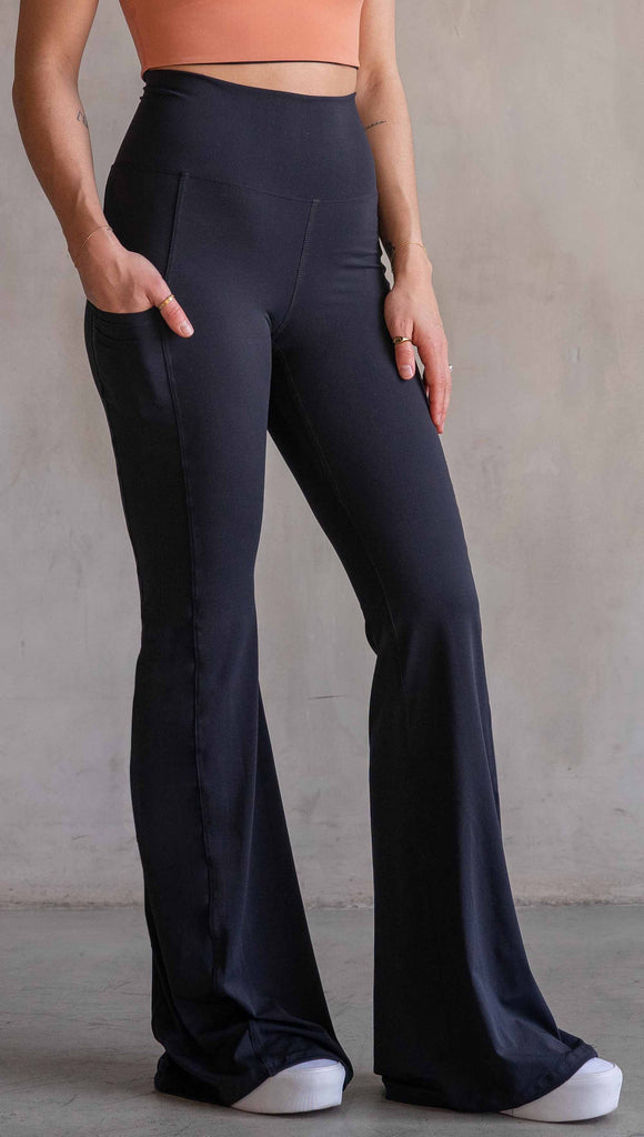 Model wearing WERKSHOP Black EnviSoft Bells. The pants feaure a belled leg opening, flatlock seams, pockets on each thigh, a long panel gusset and small silver eagle logo on the right pocket.