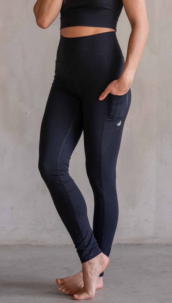 Model wearing WERKSHOP Solid Black EnviSoft Leggings. Featuring pockets on each thigh, a long panel gusset, flatlock seams and an eagle logo on the right pocket.