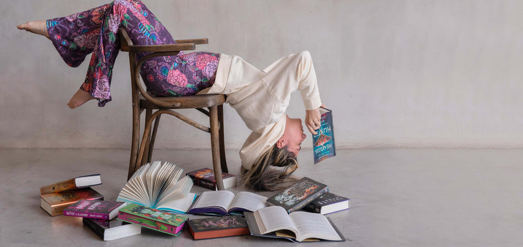 Girl doing a moderate backbend over a chair while reading her book. She is wearing WERKSHOP Enchanted Garden bells and "A Book A Day Keeps Reality Away" hoodie.