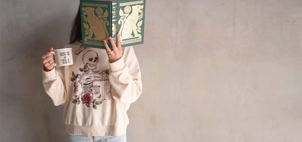 Model wearing WERKSHOP "Death by TBR" hoodie while holding up a book with one hand and a "Death by TBR" coffee mug in the other hand.