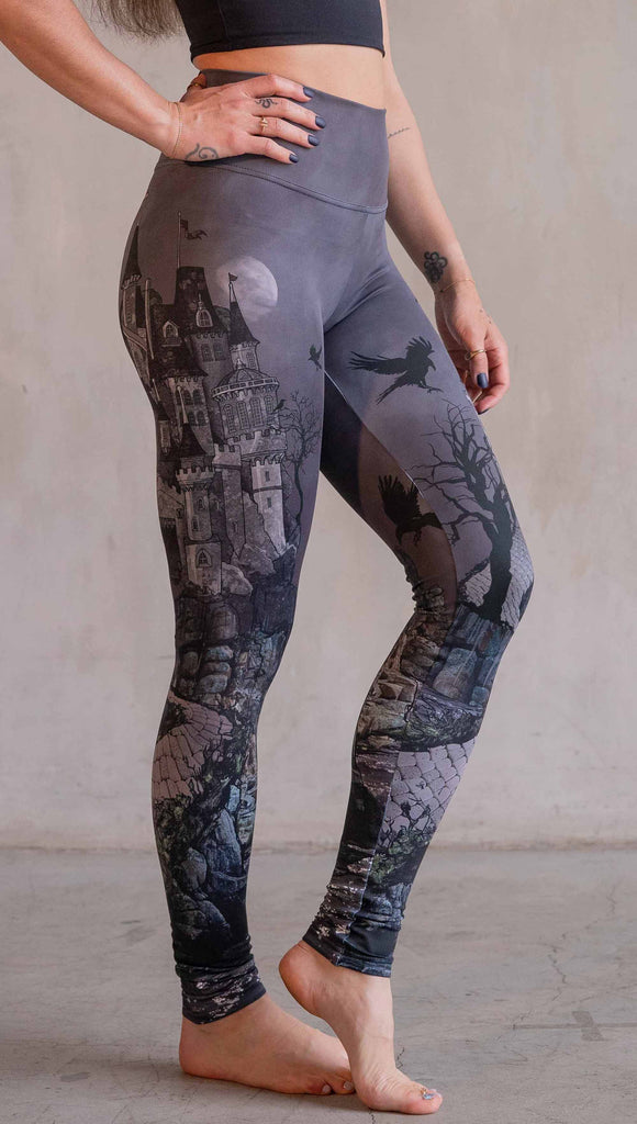 Model wearing WERKSHOP Dark Castle Leggings. The artwork on the leggings feature a fully monochrome (black and gray) gothic castle with stone walkway and crows flying in the moonlight.