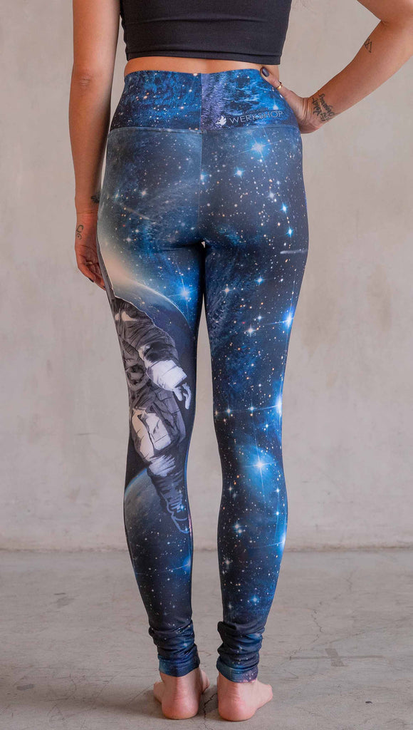 Model wearing WERKSHOP Catstronaut Leggings. The leggings are printed with a cat in an astronaut suit on the wearer's left leg. The galaxy background is a deep cobalt blue with nebula swirls and shooting stars.