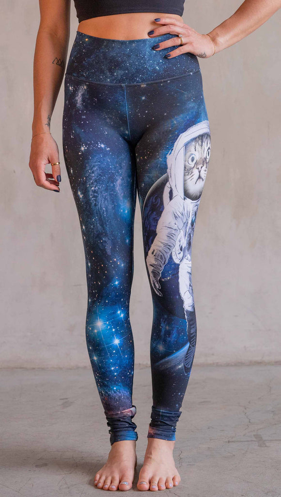 Model wearing WERKSHOP Catstronaut Leggings. The leggings are printed with a cat in an astronaut suit on the wearer's left leg. The galaxy background is a deep cobalt blue with nebula swirls and shooting stars.