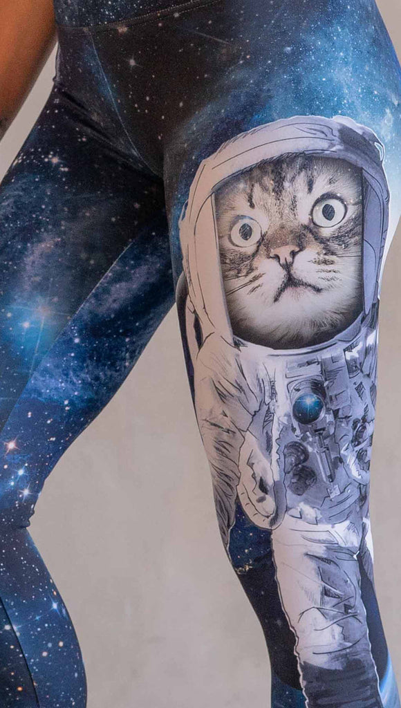 Model wearing WERKSHOP Catstronaut Leggings. The leggings are printed with a cat in an astronaut suit on the wearer's left leg. The galaxy background is a deep cobalt blue with nebula swirls and shooting stars.