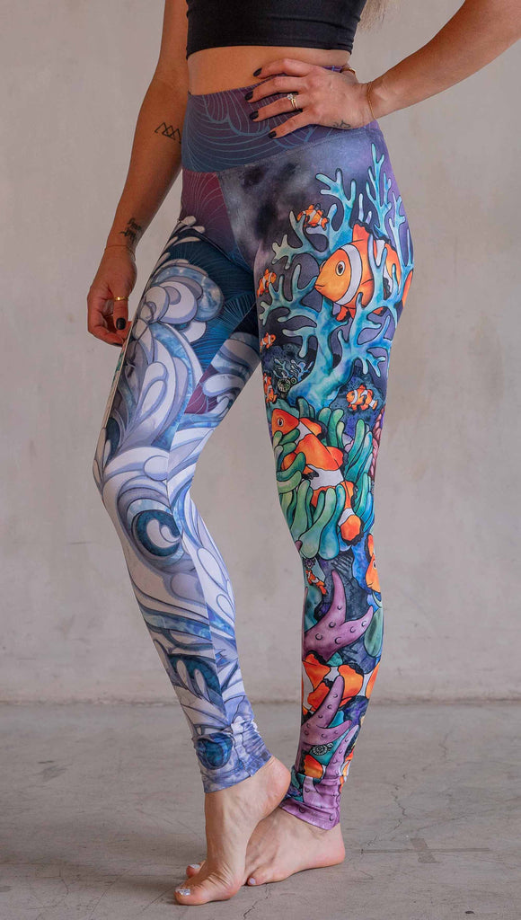 Model wearing WERKSHOP Clownfish and Surf Mashup Leggings. These leggings feature two iconic prints. on the right leg, you have a surfboard with dolphins and spashing waves. On the left leg, you have an explosion of color with a coral reef and clownfish.