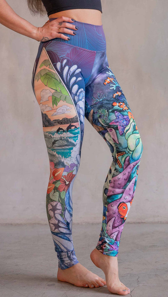 Model wearing WERKSHOP Clownfish and Surf Mashup Leggings. These leggings feature two iconic prints. on the right leg, you have a surfboard with dolphins and spashing waves. On the left leg, you have an explosion of color with a coral reef and clownfish.