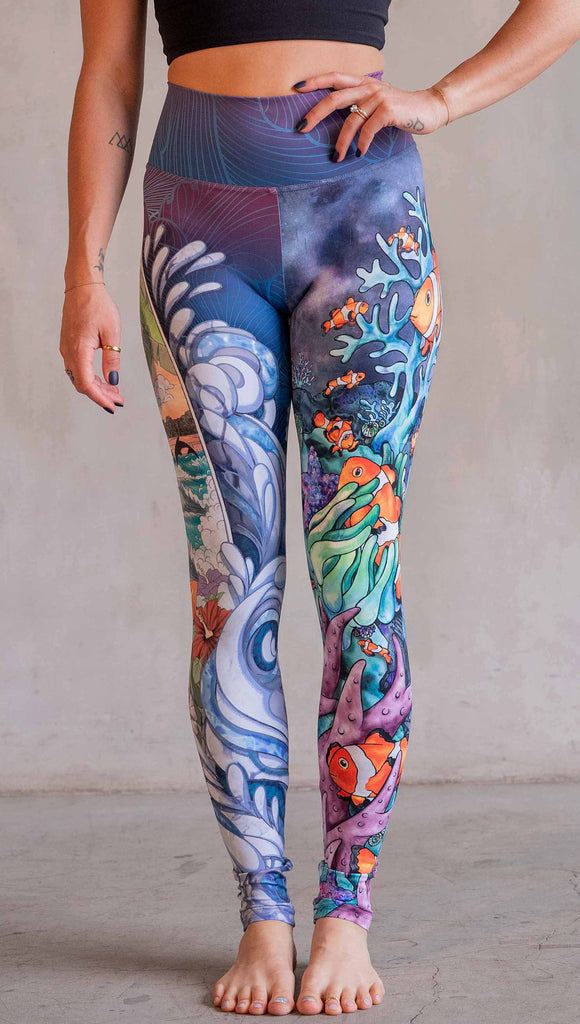 Model wearing WERKSHOP Clownfish and Surf Mashup Leggings. These leggings feature two iconic prints. on the right leg, you have a surfboard with dolphins and spashing waves. On the left leg, you have an explosion of color with a coral reef and clownfish.