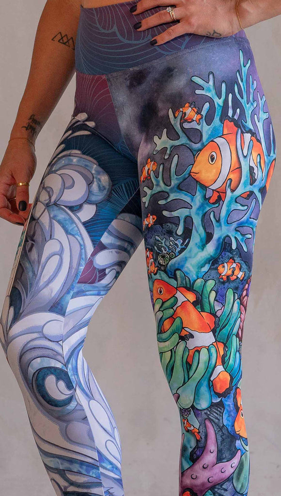 Model wearing WERKSHOP Clownfish and Surf Mashup Leggings. These leggings feature two iconic prints. on the right leg, you have a surfboard with dolphins and spashing waves. On the left leg, you have an explosion of color with a coral reef and clownfish.