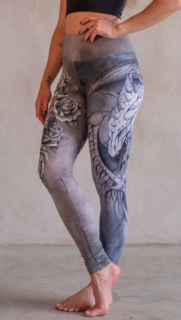 Model wearing WERKSHOP Dagger Mashup Leggings. One leg has a dagger with a wreath of roses. The other leg has an abstract gargoyle-esc dragon. Both legs are neutral in color tone with the right leg being a bit taupe and the left leg being a cool gray.