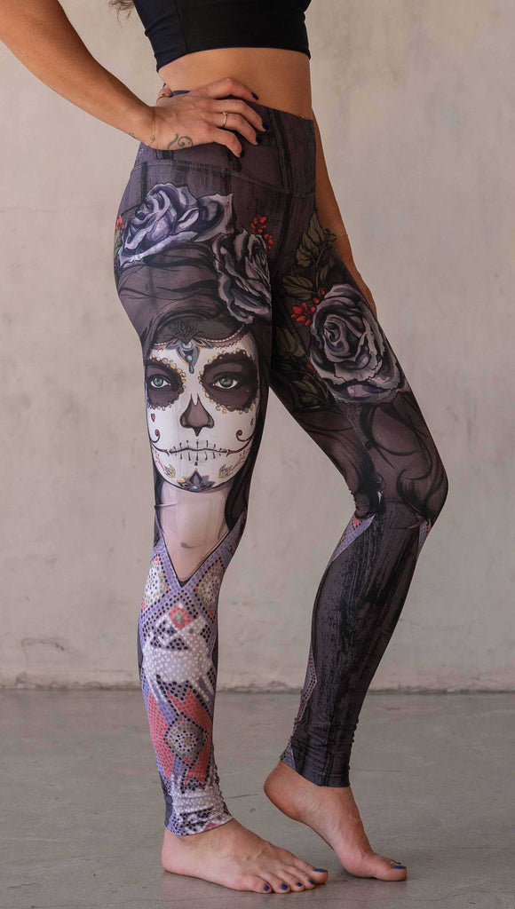 Girl wearing WERKSHOP "Dark Sugar" Athleisure leggings. The artwork on the leggings features a girl wearing dia de los muertos makeup in tones of blue and gray. she is wearing a crown of roses and a beaded necklace over a distressed warm gray woodgrain background.