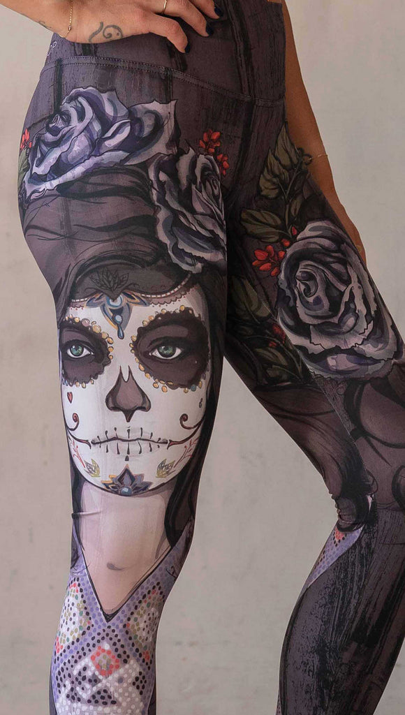 Girl wearing WERKSHOP "Dark Sugar" Athleisure leggings. The artwork on the leggings features a girl wearing dia de los muertos makeup in tones of blue and gray. she is wearing a crown of roses and a beaded necklace over a distressed warm gray woodgrain background.