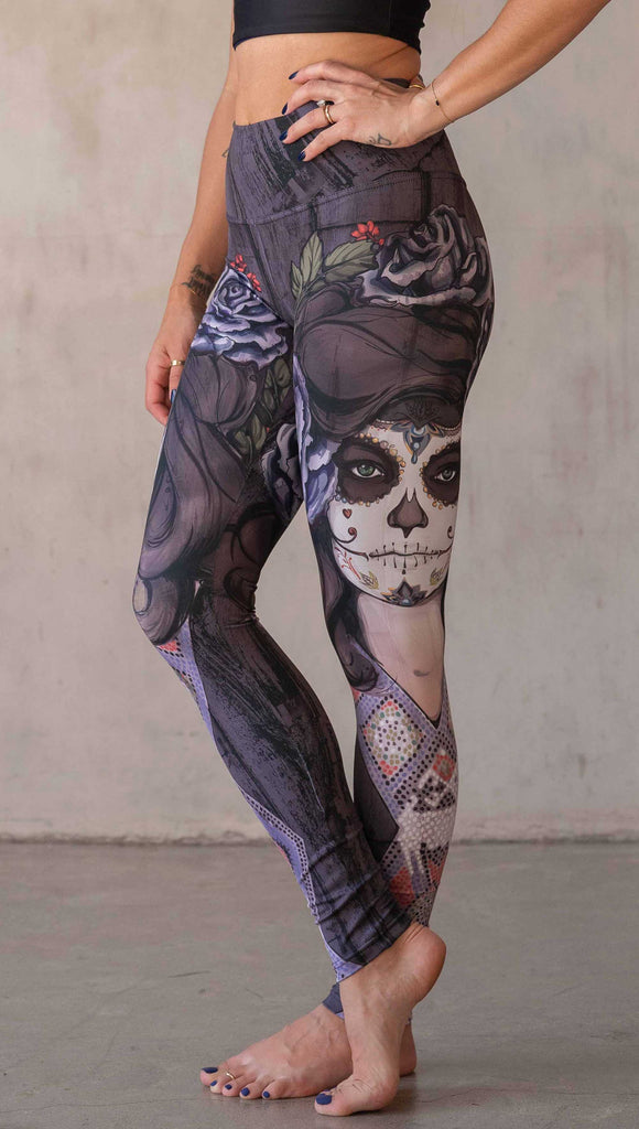 Girl wearing WERKSHOP "Dark Sugar" Athleisure leggings. The artwork on the leggings features a girl wearing dia de los muertos makeup in tones of blue and gray. she is wearing a crown of roses and a beaded necklace over a distressed warm gray woodgrain background.