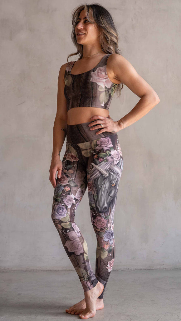 Model wearing WERKSHOP Dark Unicorn Athleisure Leggings. The leggings feature original artwork of a unicorn surrounded by a wreath of vintage coloured roses. The unicorn has a beaded chain with a skull dangling from it's horn. The artwork is over a warm woodgrain background.