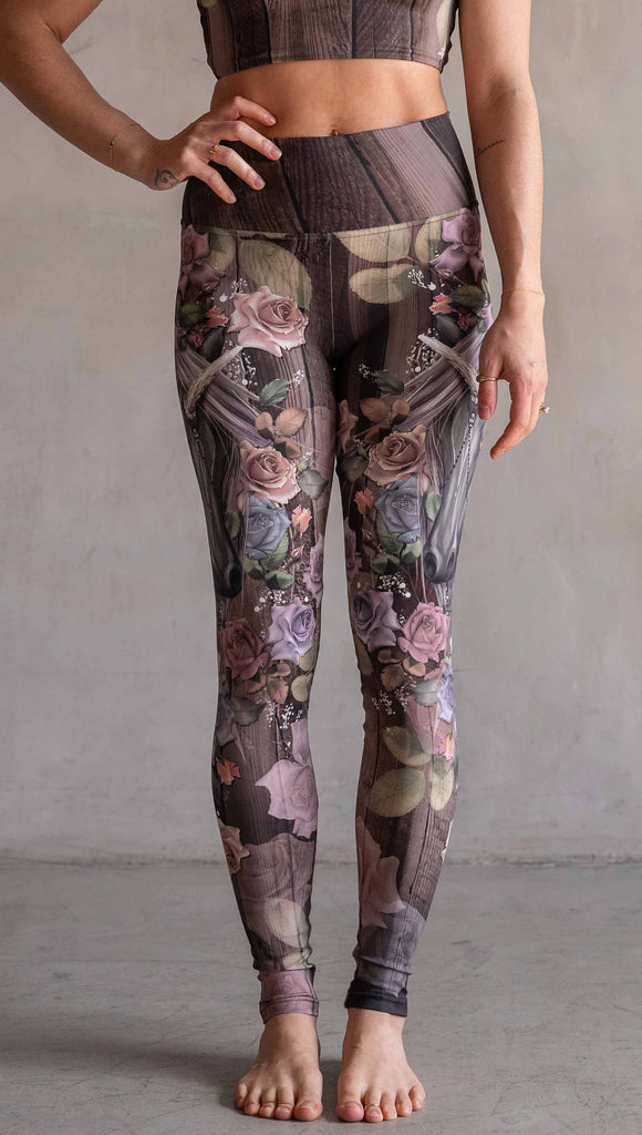 Model wearing WERKSHOP Dark Unicorn Athleisure Leggings. The leggings feature original artwork of a unicorn surrounded by a wreath of vintage coloured roses. The unicorn has a beaded chain with a skull dangling from it's horn. The artwork is over a warm woodgrain background.