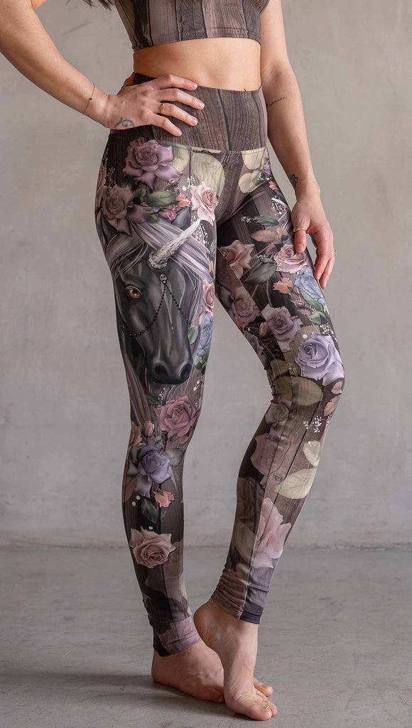 Model wearing WERKSHOP Dark Unicorn Athleisure Leggings. The leggings feature original artwork of a unicorn surrounded by a wreath of vintage coloured roses. The unicorn has a beaded chain with a skull dangling from it's horn. The artwork is over a warm woodgrain background.