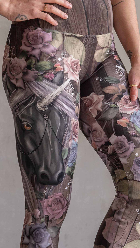 Model wearing WERKSHOP Dark Unicorn Athleisure Leggings. The leggings feature original artwork of a unicorn surrounded by a wreath of vintage coloured roses. The unicorn has a beaded chain with a skull dangling from it's horn. The artwork is over a warm woodgrain background.