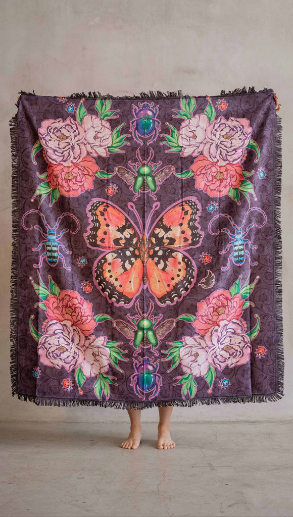 WERKSHOP Enchanted Garden tapestry draped on a couch. The tapestry is printed with original artwork by Chriztina Marie. The artwork printed not the fabric features Butterflies, Beetles and Peonies over a warm fuchsia with bright bold pops of color on each beetle and Butterfly.