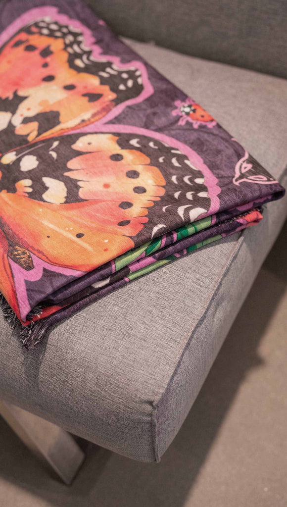 WERKSHOP Enchanted Garden tapestry draped on a couch. The tapestry is printed with original artwork by Chriztina Marie. The artwork printed not the fabric features Butterflies, Beetles and Peonies over a warm fuchsia with bright bold pops of color on each beetle and Butterfly.