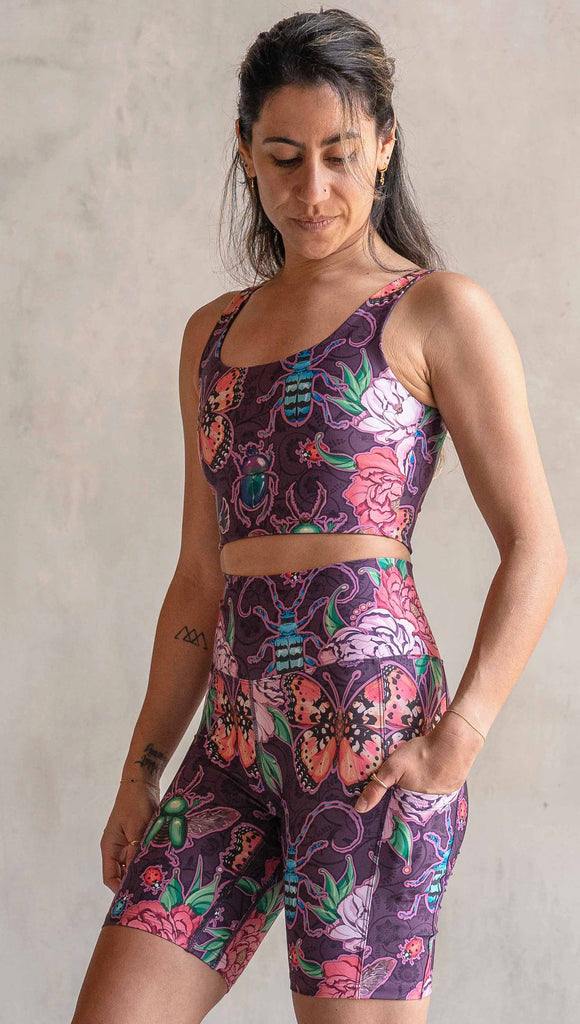 Side view of model wearing WERKSHOP Enchanted Garden EnviSoft Bicycle length shorts with pockets. The fabric is printed with original artwork by our Female Founder, Chriztina Marie. The artwork printed not the fabric features Butterflies, Beetles and Peonies over a warm fuchsia with bright bold pops of color on each beetle and Butterfly.