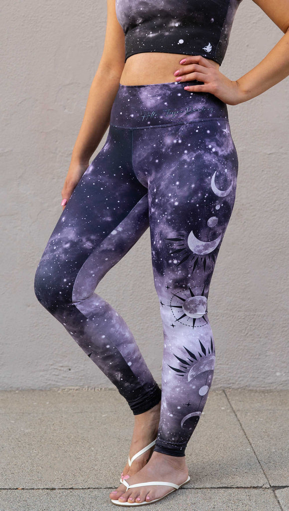 Model wearing "Feyre's Tattoo" Leggings with a dark purplish-blue to black ombre celestial background and moon phases on each leg. Officially licensed for A Court of Thorns and Roses by Sarah J Maas. It has the phrase, "Hello, Feyre Darling" on the waistband.