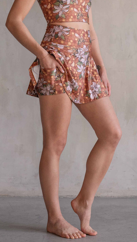 Model wearing WERKSHOP Featherlight set with Honeybess Skirt and Top. The skirt features built-in shorts with pockets. The fabric is printed with original artwork by Chriztina Marie that features clusters of honeybees and flowers over a gold honeycomb inspired background. The skirt falls to about the wearer's fingertips and the shorts are hidden underneath.