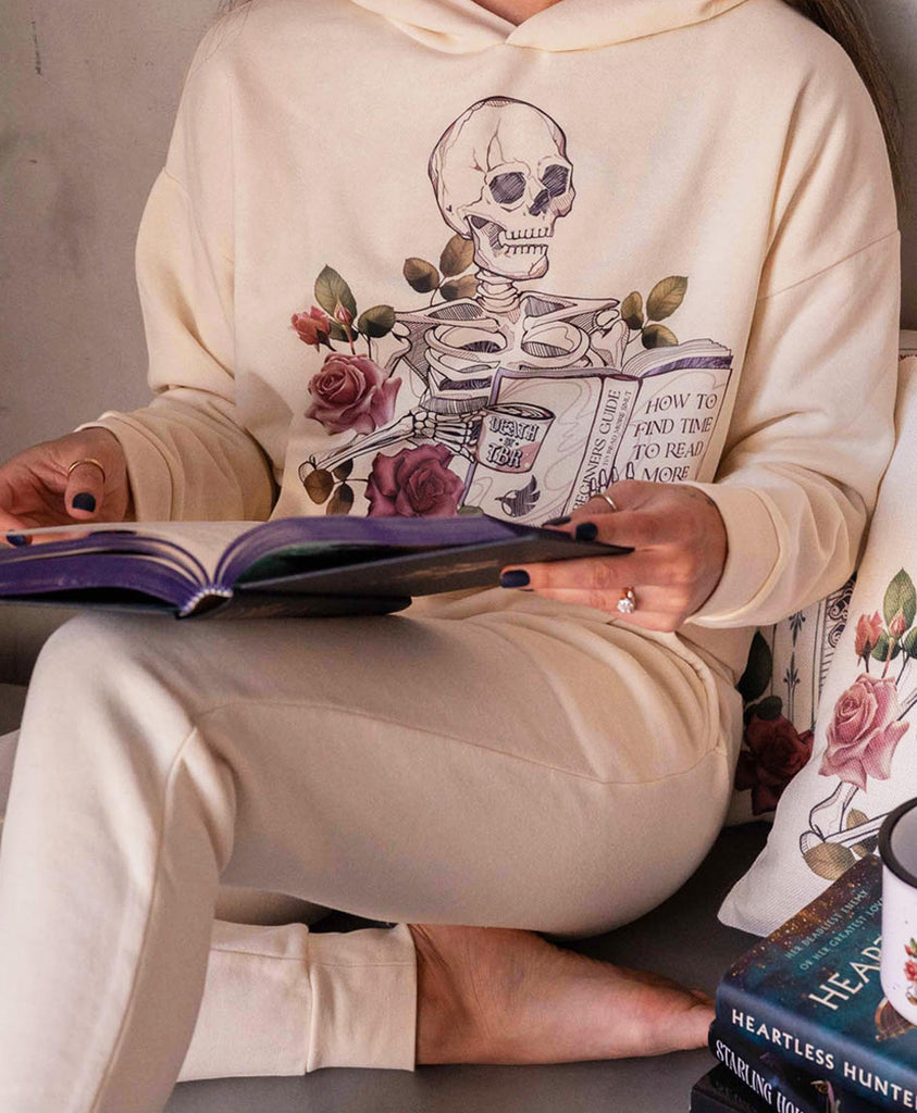 Girl casually sitting on the floor while reading a book. She is wearing WERKSHOP "Death by TBR" hoodie and jogger set in Bone Color.