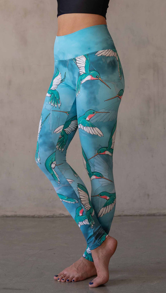 Side view of model wearing WERKSHOP Hummingbirds athleisure leggings. The artwork on the leggings fetures hummingbirds fluttering through the sky over an aqua watercolor background