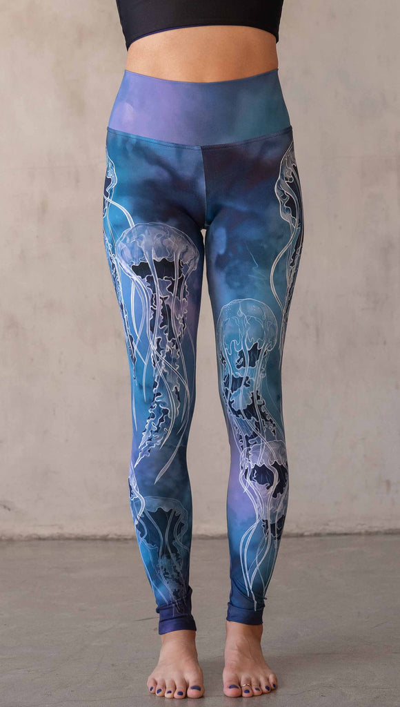 Model wearing WERKSHOP Jellyfish Athleisure Leggings. The artwork on the leggings shows white jellyfish swimming up the wearer's legs over a cobalt blue and purple watercolor background.