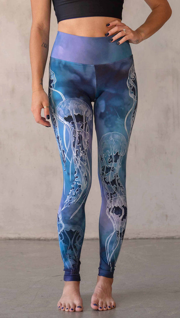 Model wearing WERKSHOP Jellyfish Athleisure Leggings. The artwork on the leggings shows white jellyfish swimming up the wearer's legs over a cobalt blue and purple watercolor background.