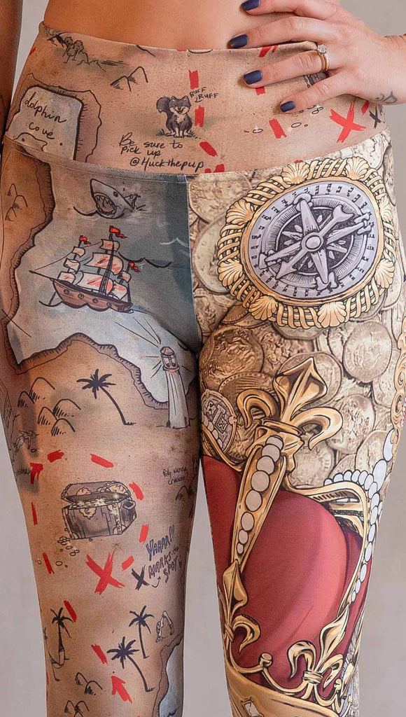 Model wearing WERKSHOP Booty Mashup Leggings. The wearer's right leg is printed with vintage inspired, hand drawn, treasure map artwork and the left leg is golden treasure with a kings royal crown, keys and pearls: everything you'd expect to find inside a pirates treasure.