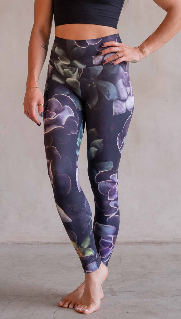 Model wearing WERKSHOP Midnight Garden Athleisure Leggings. The artwork on the leggings is printed with succulents in dark romantic colors of purple with small pops of teal