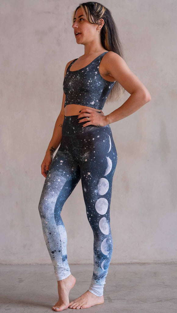 Model wearing WERKSHOP Moon Phases Athleisure Leggings. The leggings feature the phases of the moon down the wearer's left leg over a starry blue galactic background. All 12 zodiac constellations are sprinkled throughout the background of the design.