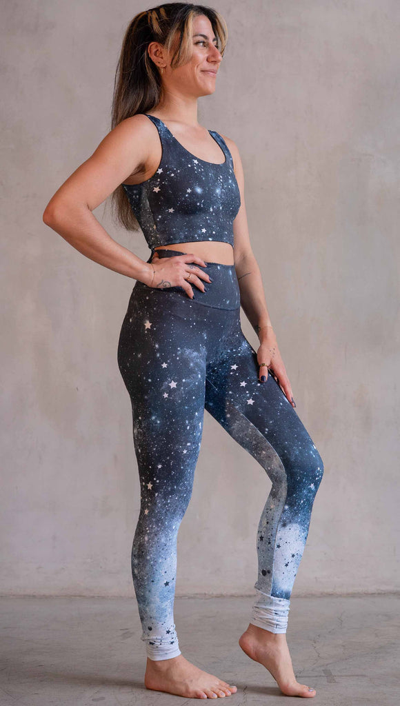 Model wearing WERKSHOP Moon Phases Athleisure Leggings. The leggings feature the phases of the moon down the wearer's left leg over a starry blue galactic background. All 12 zodiac constellations are sprinkled throughout the background of the design.