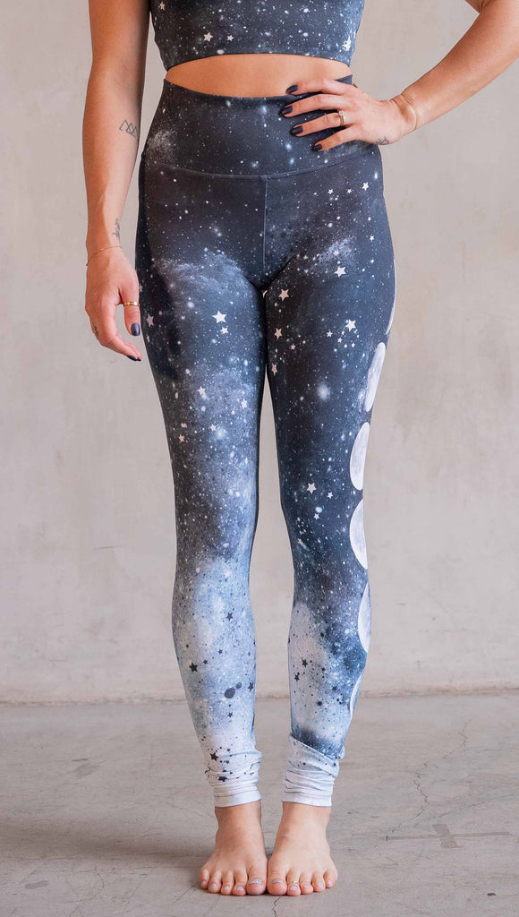 Model wearing WERKSHOP Moon Phases Athleisure Leggings. The leggings feature the phases of the moon down the wearer's left leg over a starry blue galactic background. All 12 zodiac constellations are sprinkled throughout the background of the design.