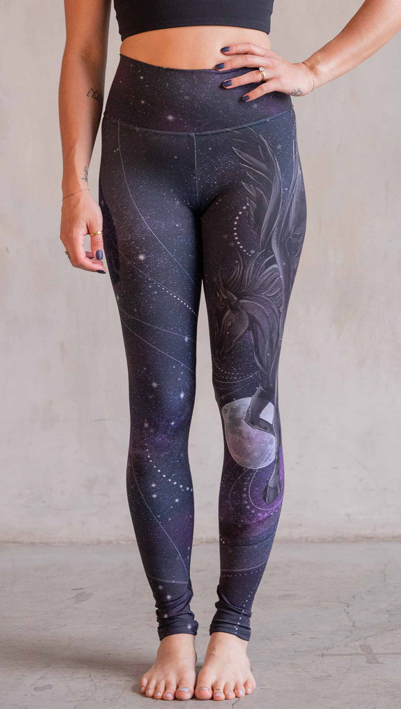 Model wearing WERKSHOP Nightmare Athleisure leggings. The artwork on the leggings features a pegasus flying through a purple galactic sky on the wearers left leg with swirls and a full moon just below the left knee.