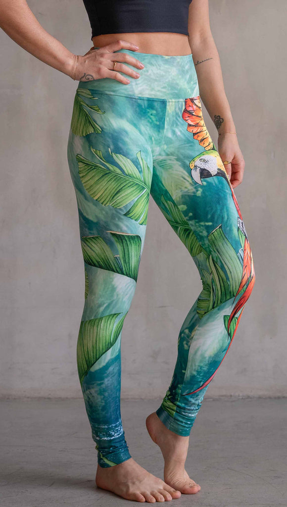 Model wearing WERKSHOP Parrot Athleisure Leggings. The artwork on the leggings feature a tropical parrot down the wearer's left leg and bright green and teal floral artwork on the right leg.